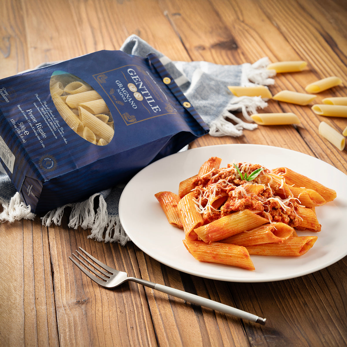 Penne rigate pasta and GENTILE bag on wooden table with plated pasta