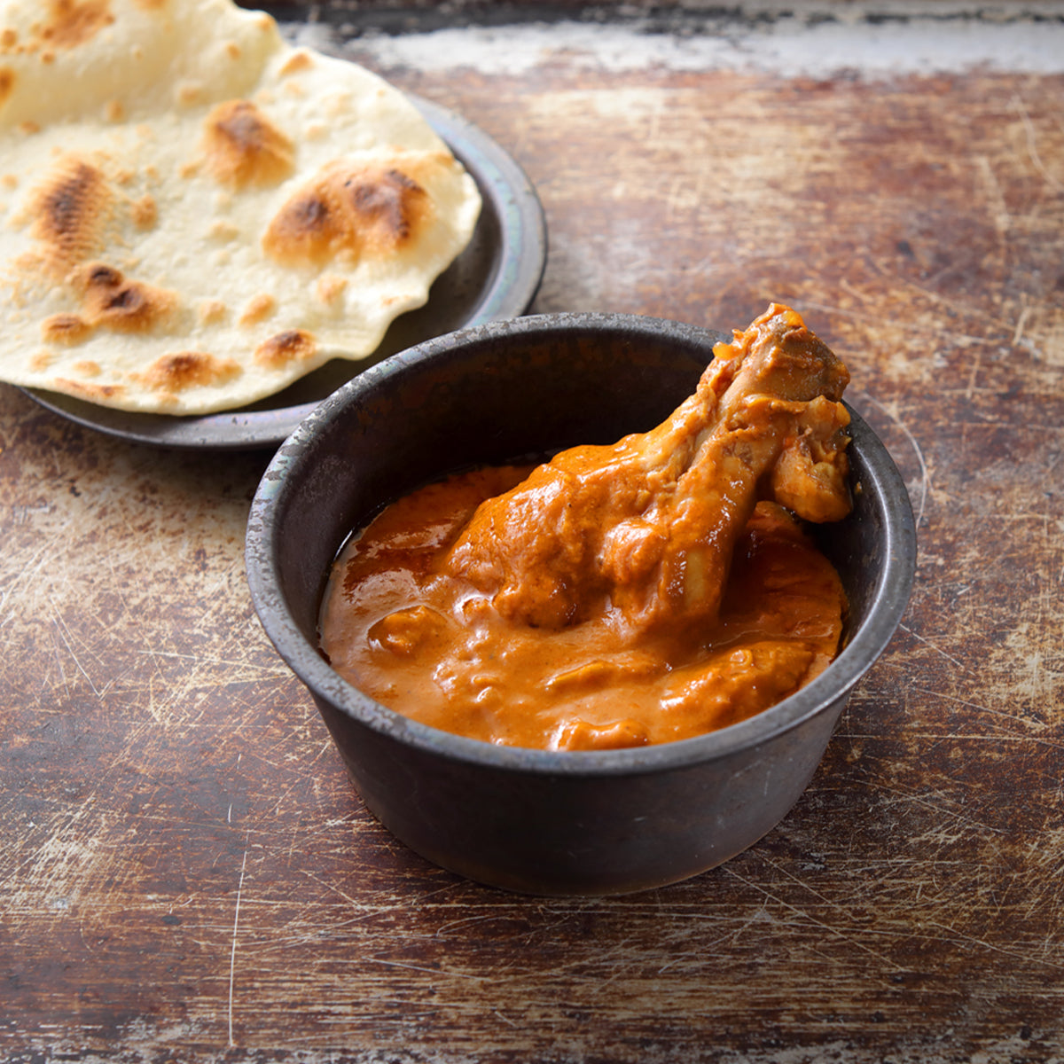 Bowl of orange butter chicken curry with chicken pieces and naan breads