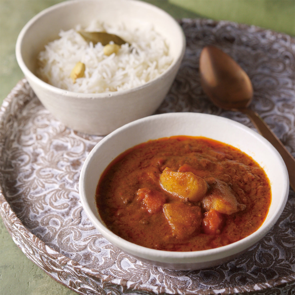 Spicy tomato curry in bowl with rice bowl and serving spoon