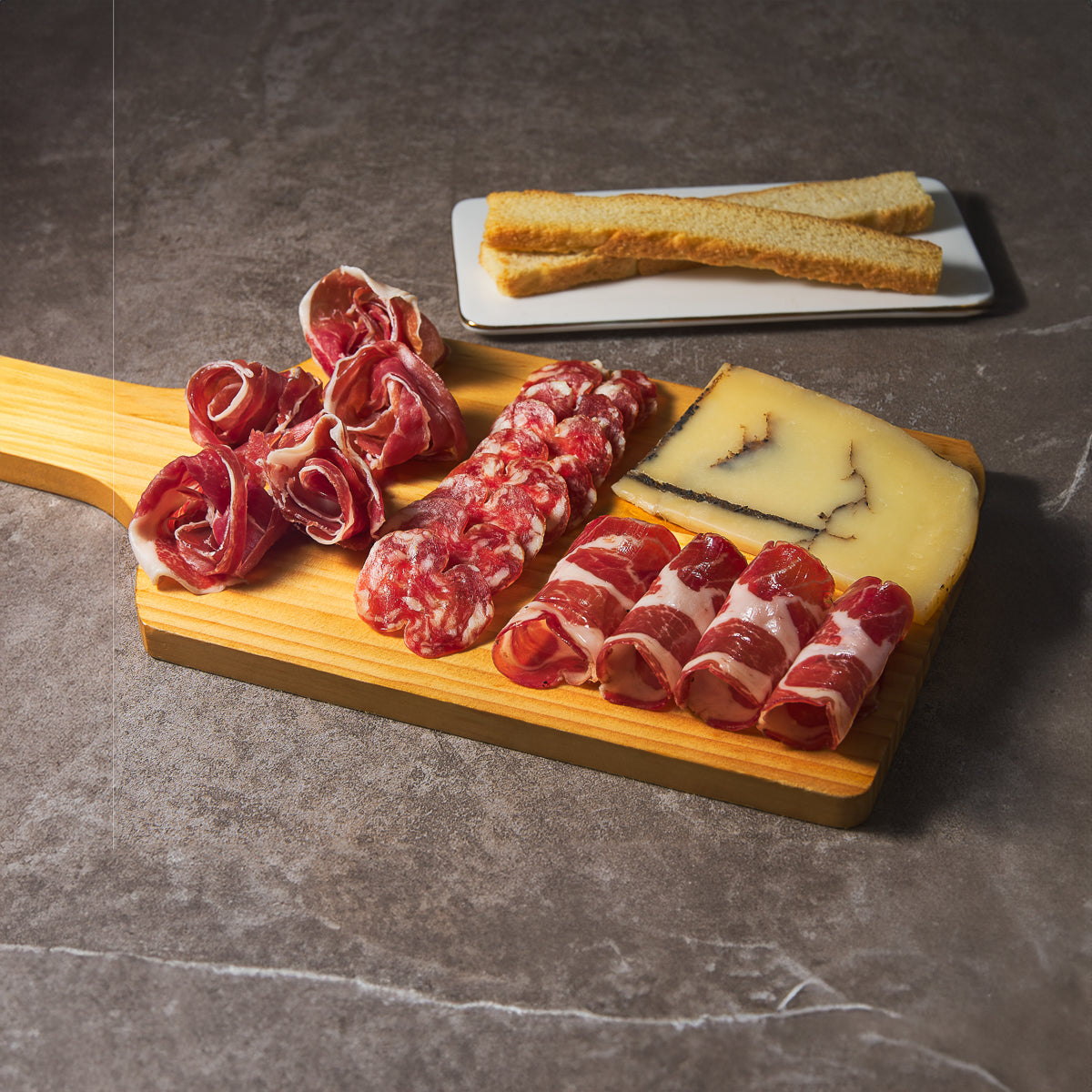 Wood deli board with sliced cured meats, cheese wedges, and bread slices on side