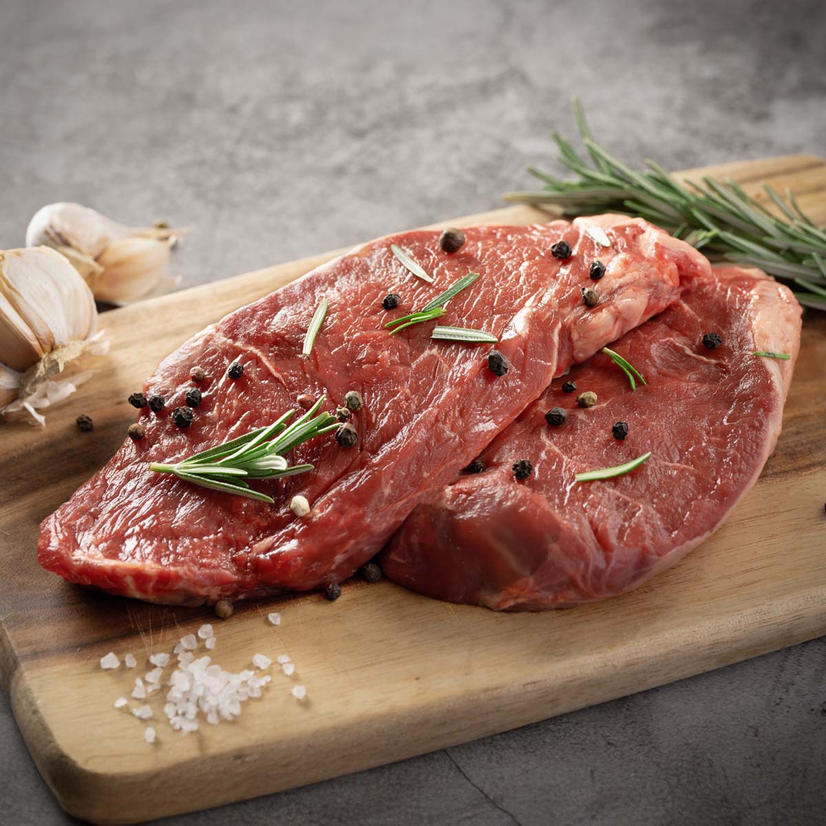 Organic beef steaks with rosemary, pepper, and sliced garlic on a wooden board